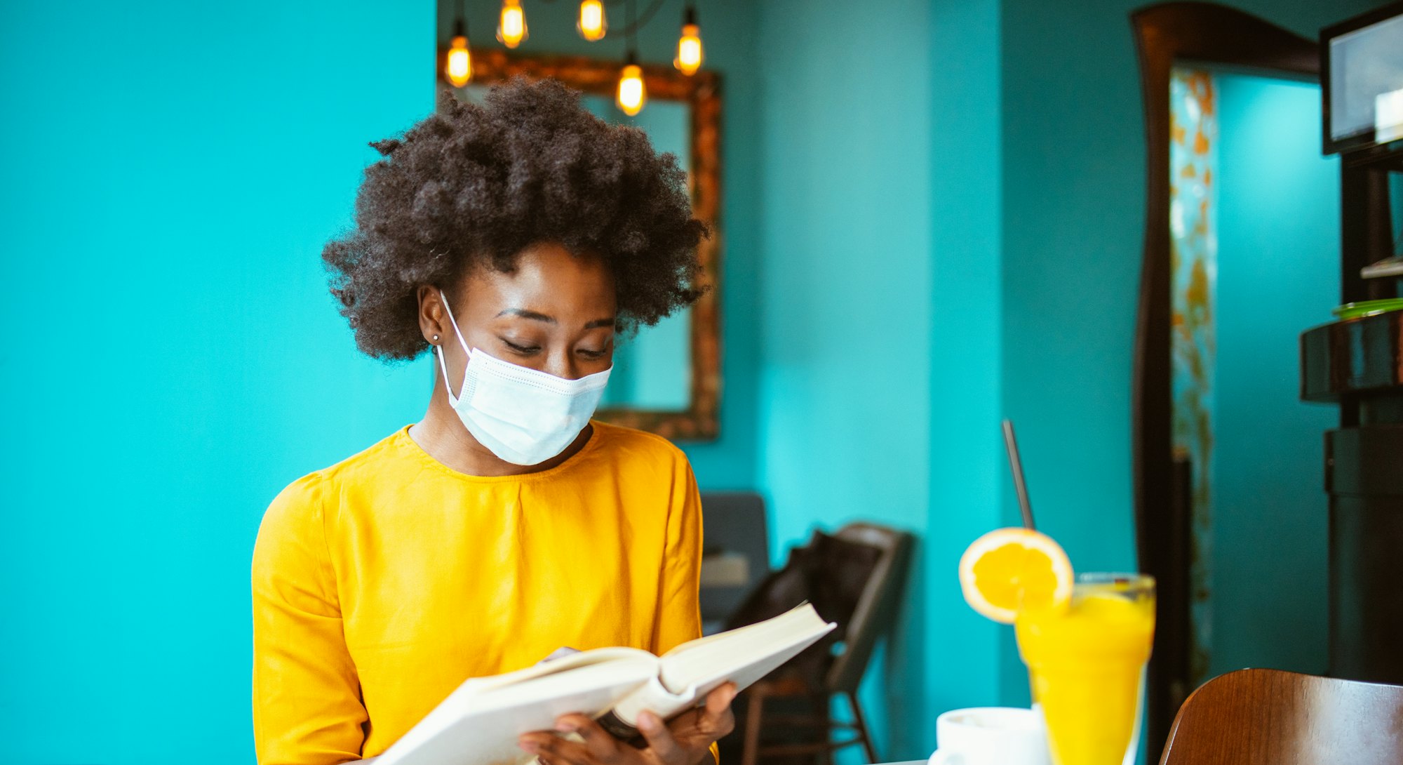 Young African-American woman enjoying reading a book and wearing protective facemask in a coffee sho...