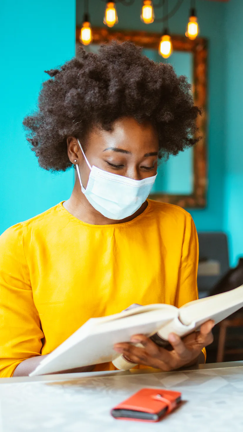 Young African-American woman enjoying reading a book and wearing protective facemask in a coffee sho...