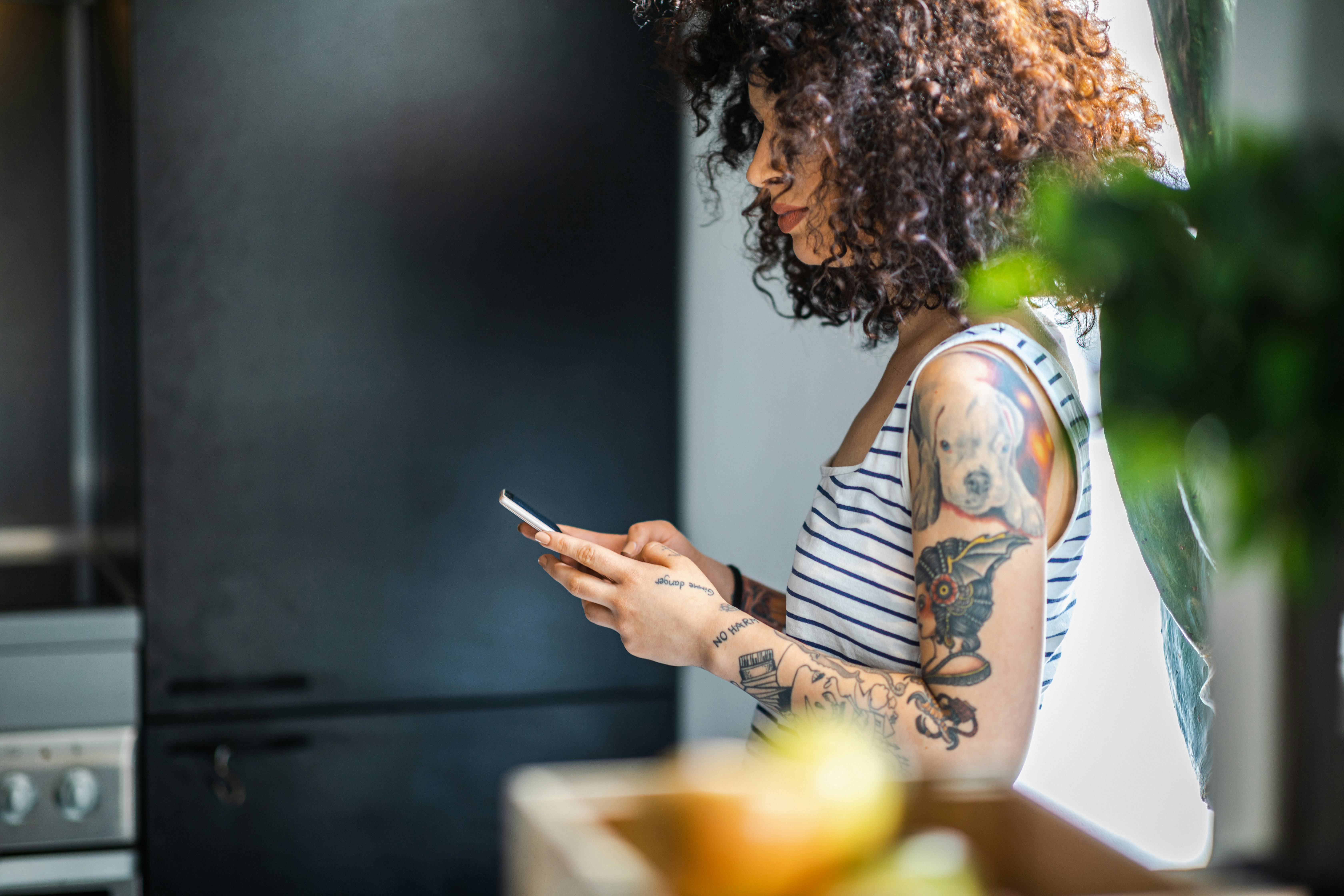 Tattooed young woman with curly hair wearing sleeveless top standing in the kitchen and using smart ...
