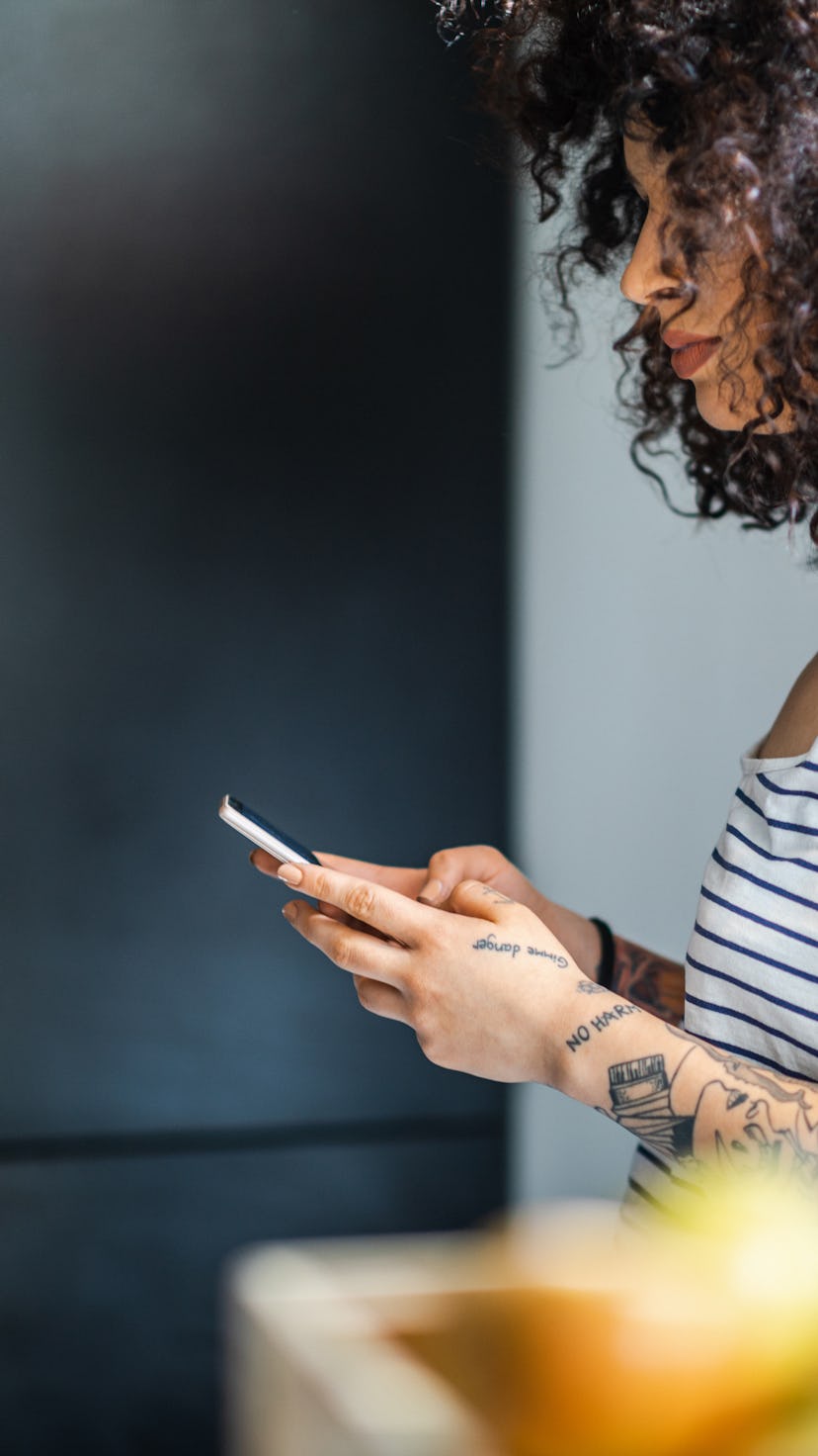 Tattooed young woman with curly hair wearing sleeveless top standing in the kitchen and using smart ...