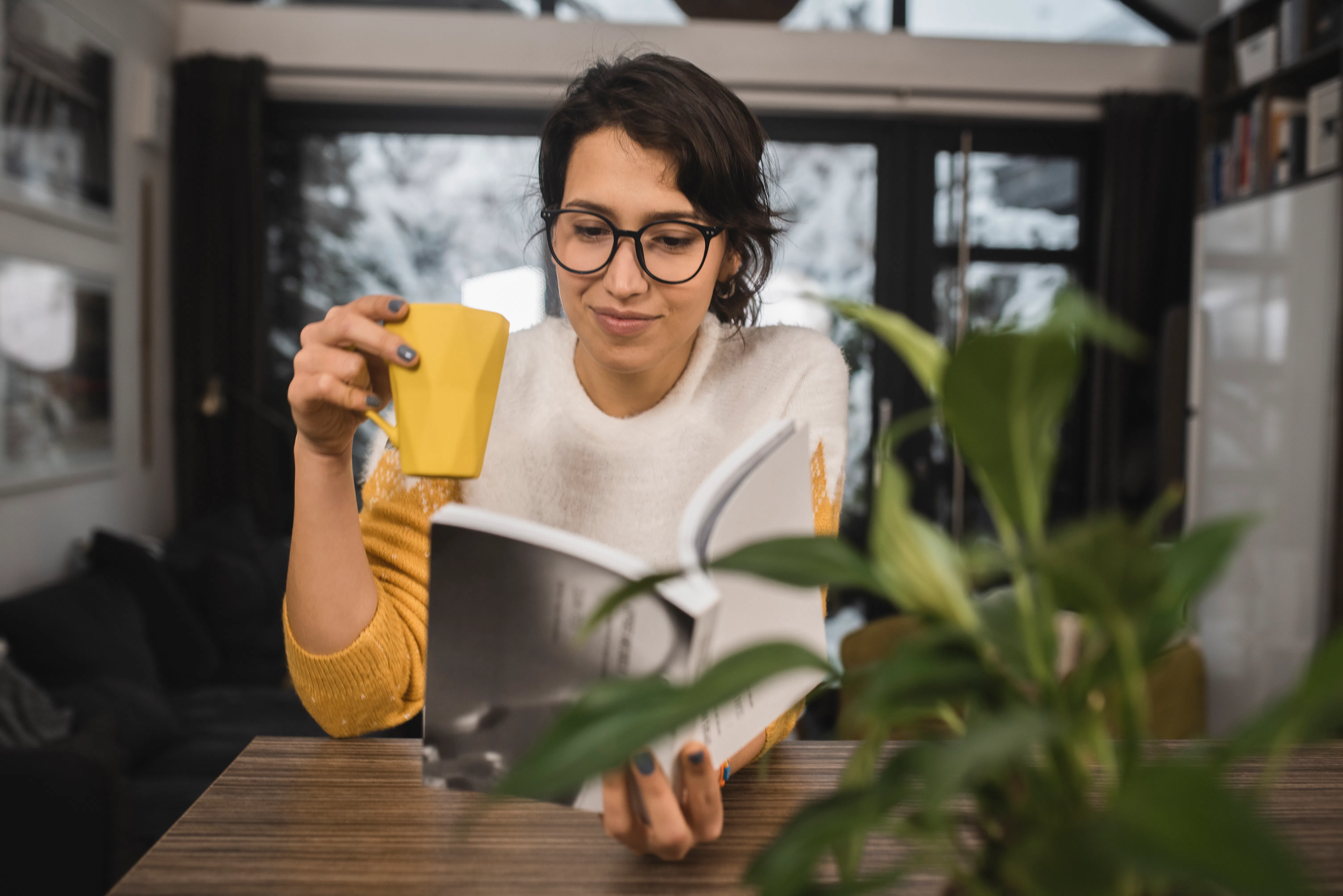 Portrait of a young woman reading a book and enjoying coffee at home.