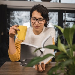 Portrait of a young woman reading a book and enjoying coffee at home.