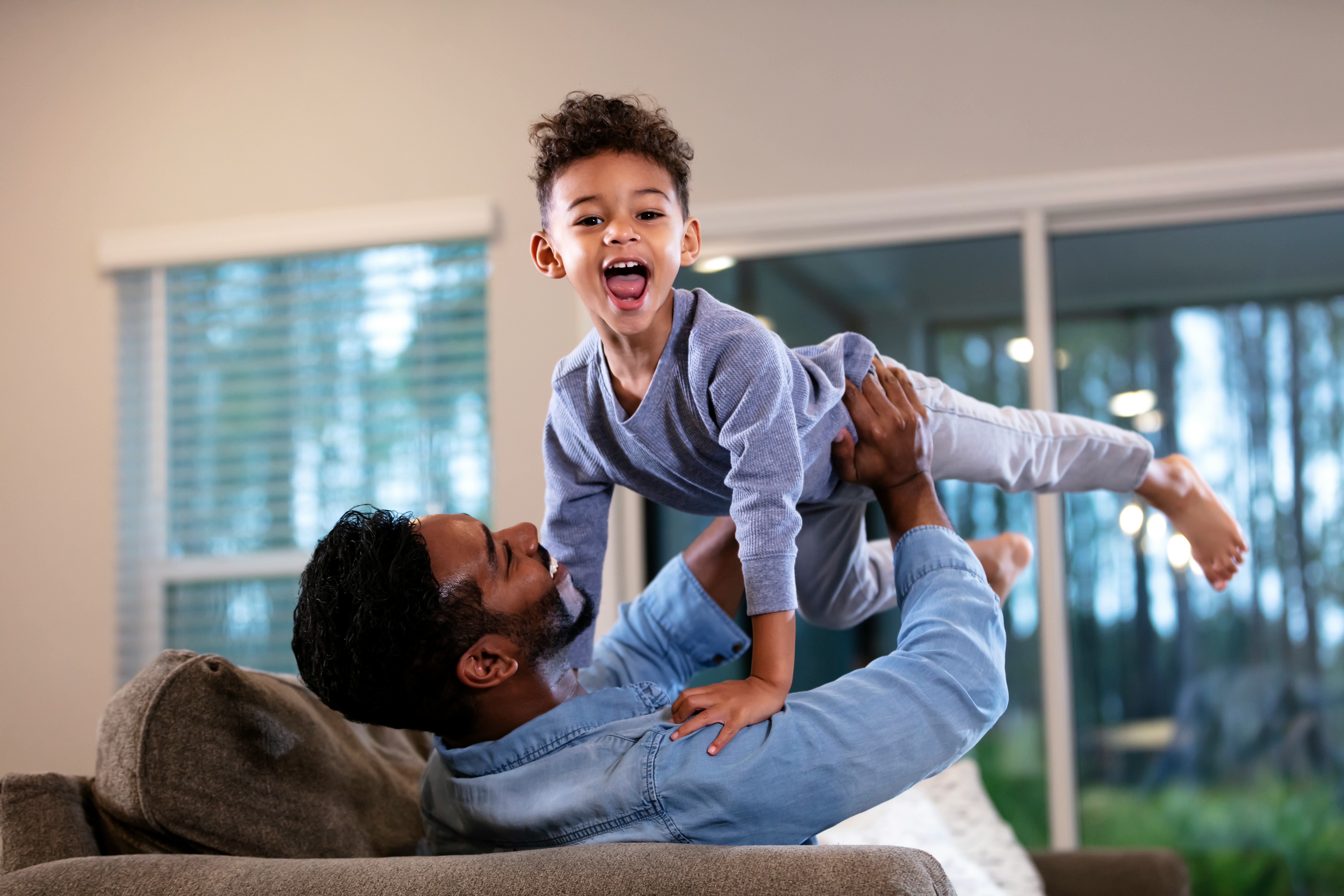 A father picks up his son, holding him aloft. The boy is laughing and shouting, mouth wide open, loo...