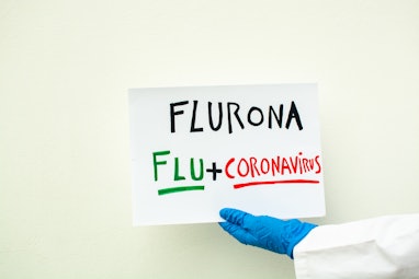 hand in lab coat and blue surgical glove holding a sign with the text fluron, coranavirus + flu, cov...