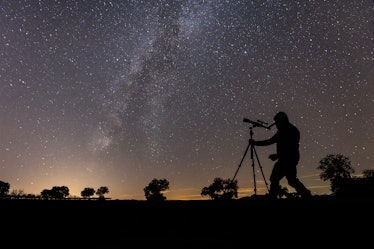 Silhouette of man with a telescope observes the starry sky during a clear night. In the background t...
