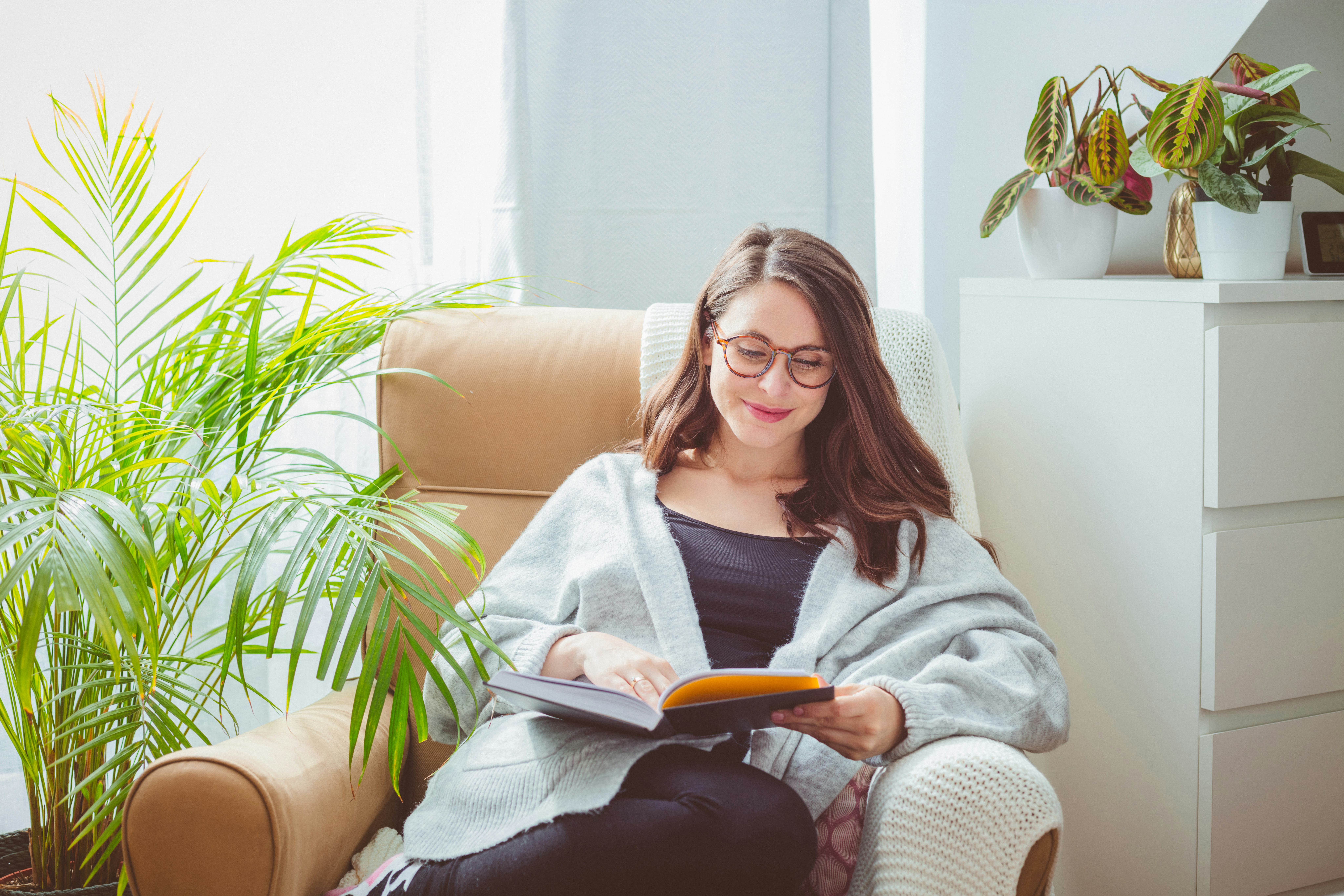 Beautiful woman wearing grey cardigan, sitting in armchair in living room and reading book.