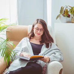 Beautiful woman wearing grey cardigan, sitting in armchair in living room and reading book.