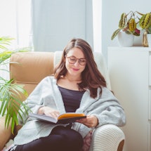 Beautiful woman wearing grey cardigan, sitting in armchair in living room and reading book.