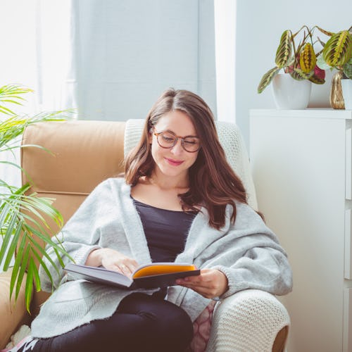Beautiful woman wearing grey cardigan, sitting in armchair in living room and reading book.