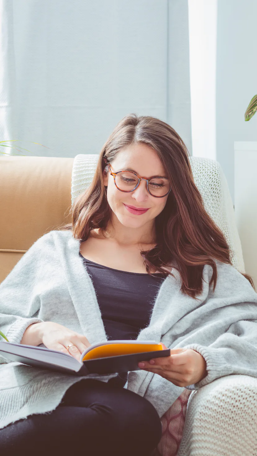 Beautiful woman wearing grey cardigan, sitting in armchair in living room and reading book.