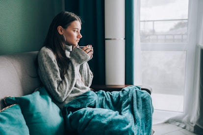 A person holding a mug, with a blanket draped over their lap, looking out a window, wondering how lo...