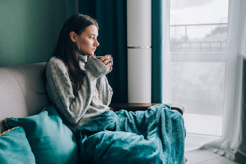 A person holding a mug, with a blanket draped over their lap, looking out a window, wondering how lo...