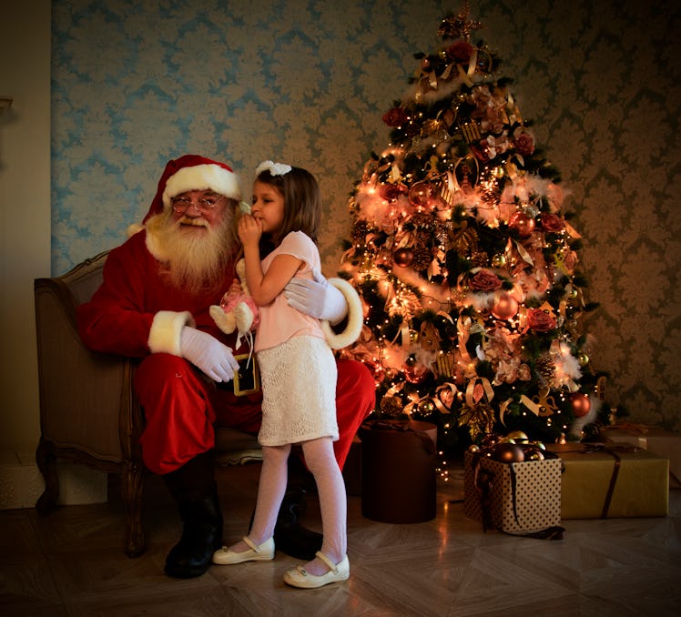 A child stands beside Santa with a Christmas tree in the background.