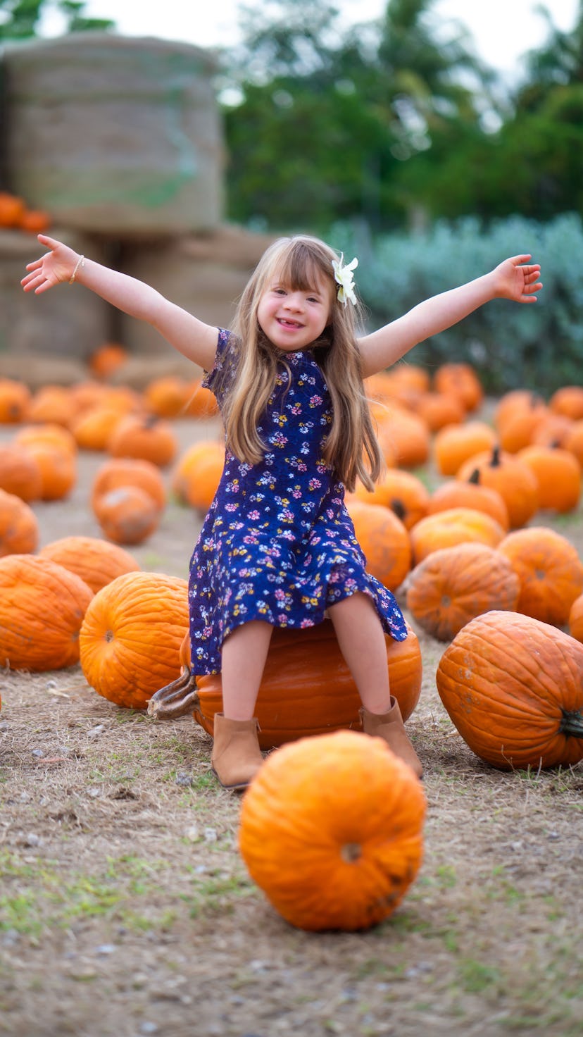 A little girl sits on a pumpkin in a pumpkin patch with her arms raised, excited about pumpkin patch...