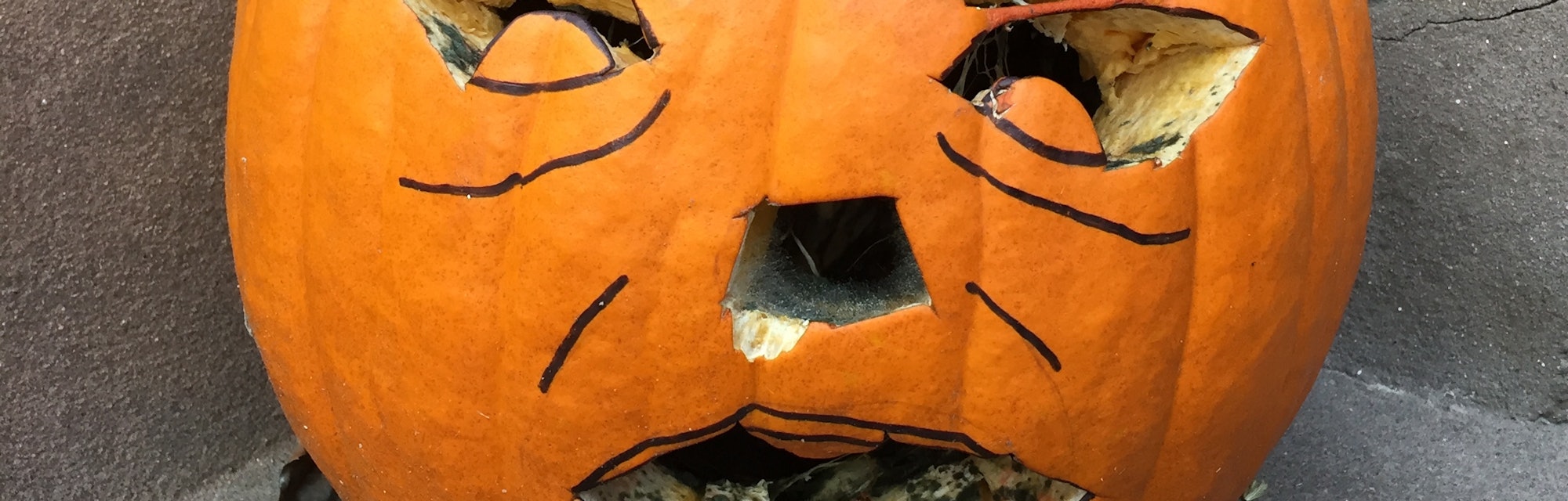 A pumpkin sitting on an outdoors brownstone wall with a carved face and rotten moldy teeth in Fort G...