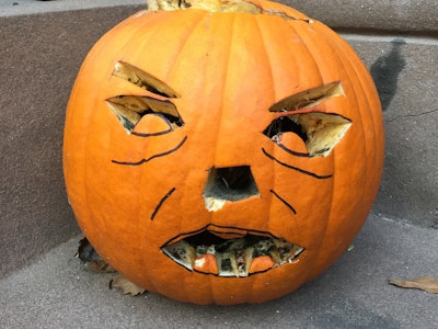 A pumpkin sitting on an outdoors brownstone wall with a carved face and rotten moldy teeth in Fort G...