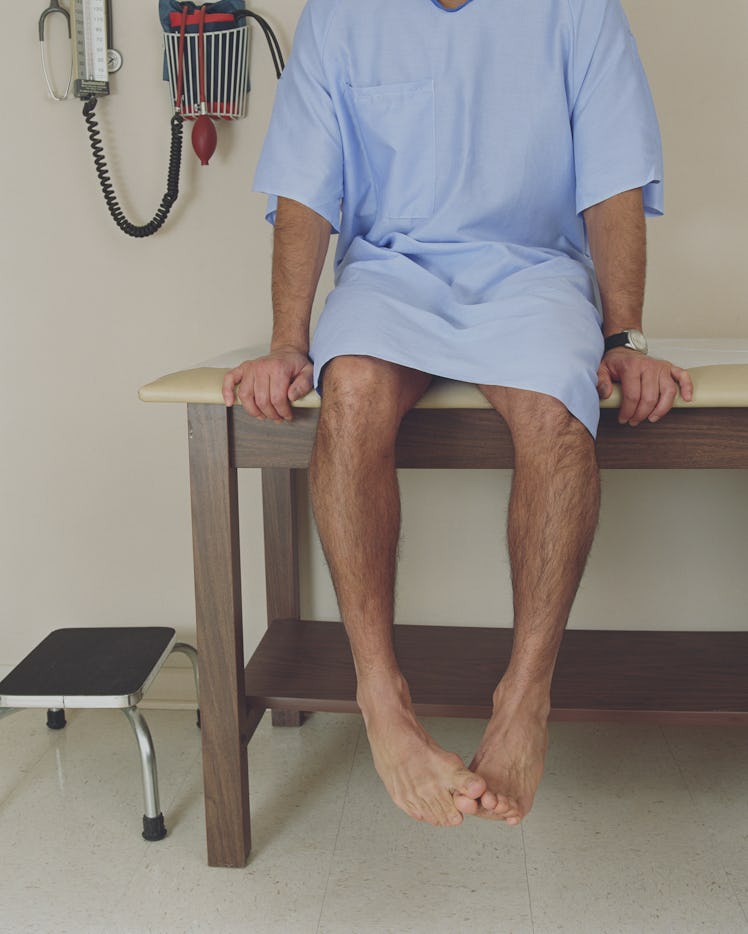 A man sits on a doctor's table in a hospital gown, He toes his curls together. His body language loo...