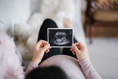 a pregnant woman lying on sofa at home, looking at the ultrasound scan photo of her baby in an artic...