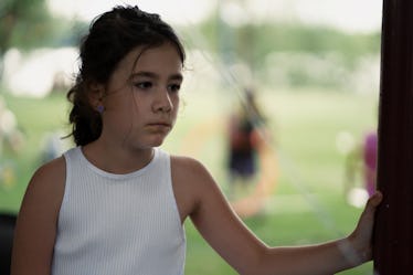 Young girl in white dress felling sad, withdrawing socially with other kids playing in the yard behi...