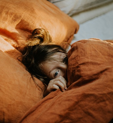 A cute young girl lies in a comfy double bed, wrapped in a thick duvet with a terracotta coloured li...