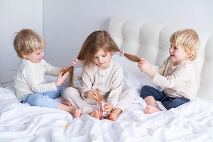 Two brothers playing with their sister's long hair while sitting on a bed.