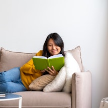Intelligent asian female lying on couch and reading book. Taking technology break and limiting scree...