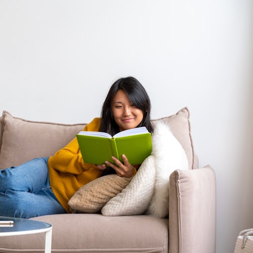 Intelligent asian female lying on couch and reading book. Taking technology break and limiting scree...