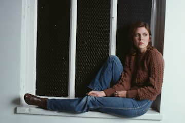 Portrait of American actress Carrie Fisher, in jeans and a brown sweater, as she sits on a window le...