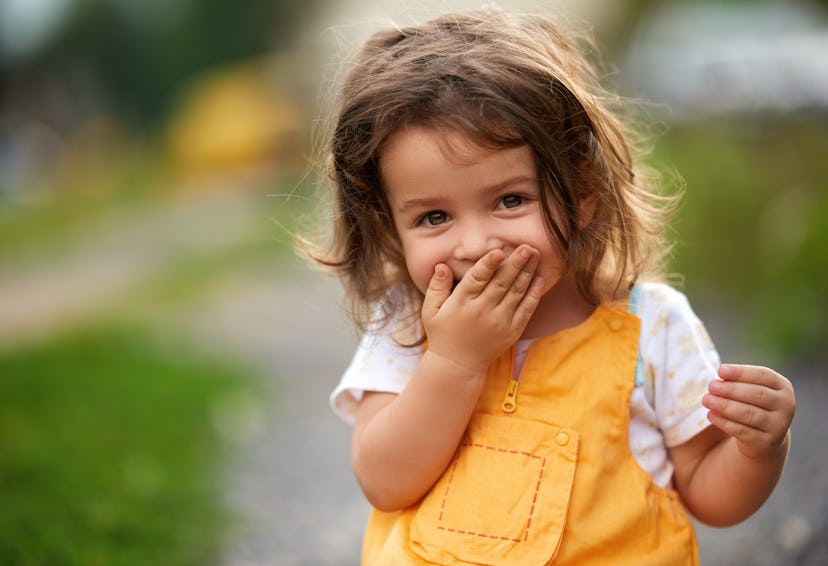 Little girl with brown hair outdoors, covering her mouth laughing in a round up of baby girl names t...
