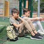 Two students with backpacks sitting together against chain link fence.