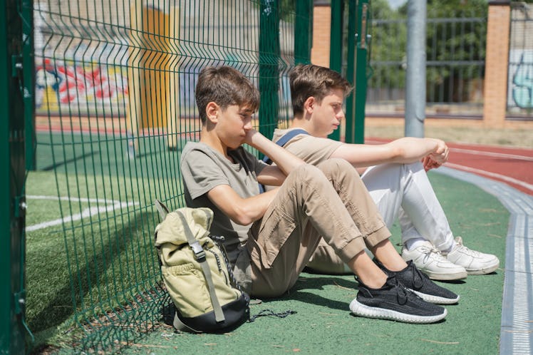 Two students with backpacks sitting together against chain link fence.