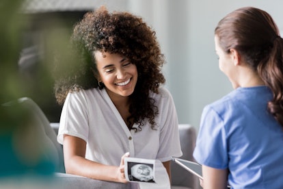Young mixed race woman smiles while looking at her baby's ultrasound image. She is meeting with a ho...