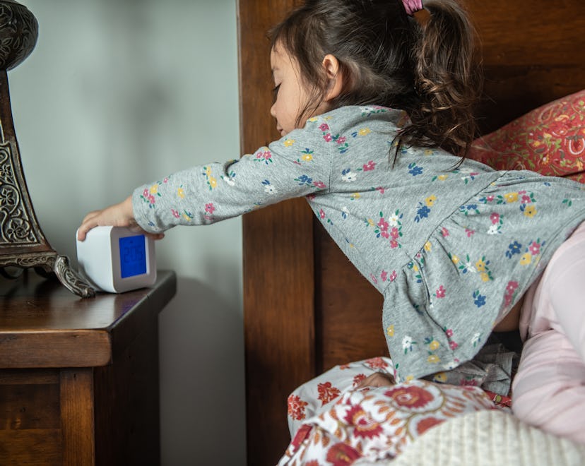A toddler girl leans across the bed to hit the snooze button on the alarm clock.