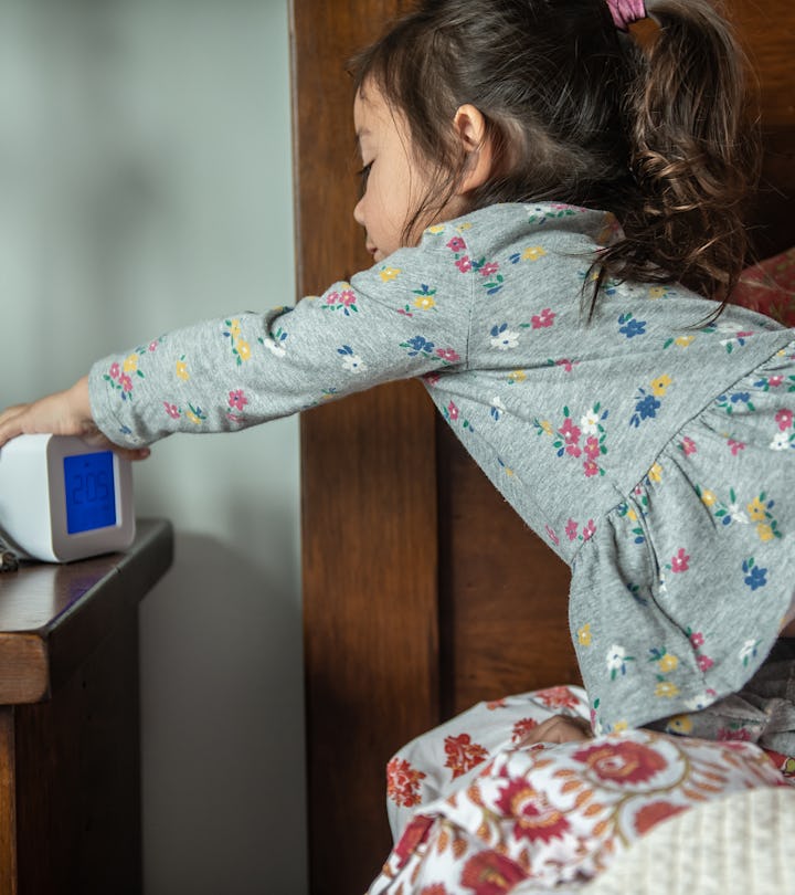 A toddler girl leans across the bed to hit the snooze button on the alarm clock.