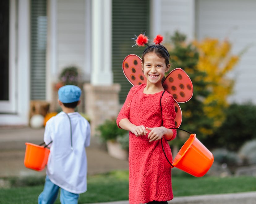 A young girl smiling in a red dress with wings, holding a treat bucket, wearing a last-minute Hallow...
