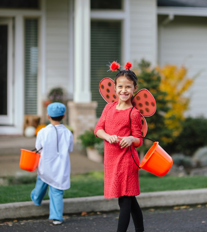 A young girl smiling in a red dress with wings, holding a treat bucket, wearing a last-minute Hallow...