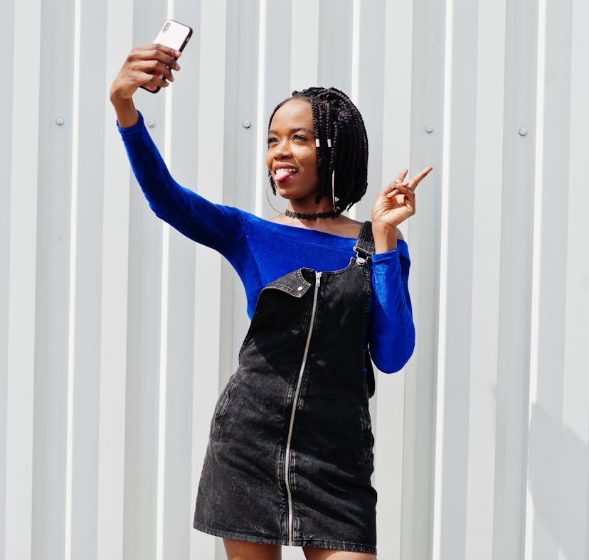 African woman in denim overalls skirt and blue dress posed against white steel wall and making selfi...