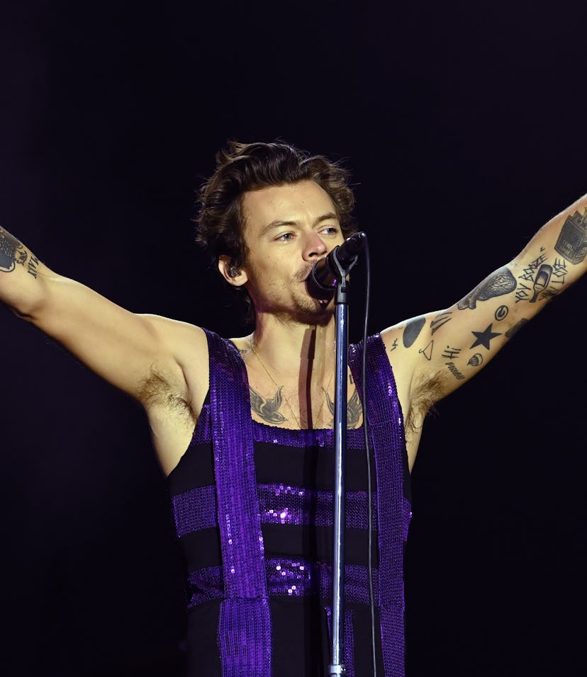 Harry Styles performs on stage with his hands in the air wearing a purple and black outfit.