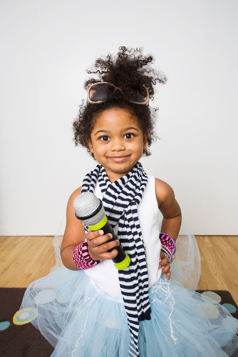 A child holding a toy microphone, wearing a striped scarf, fluffy skirt, sunglasses on her head, and...