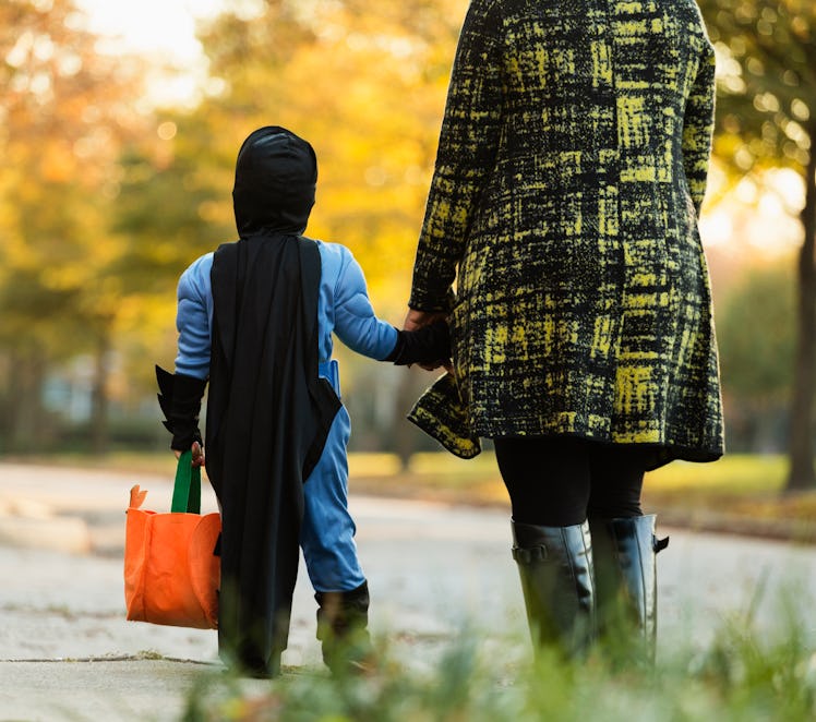 A kid wearing a batman costume and carrying an orange bag holds his moms hand while trick or treatin...
