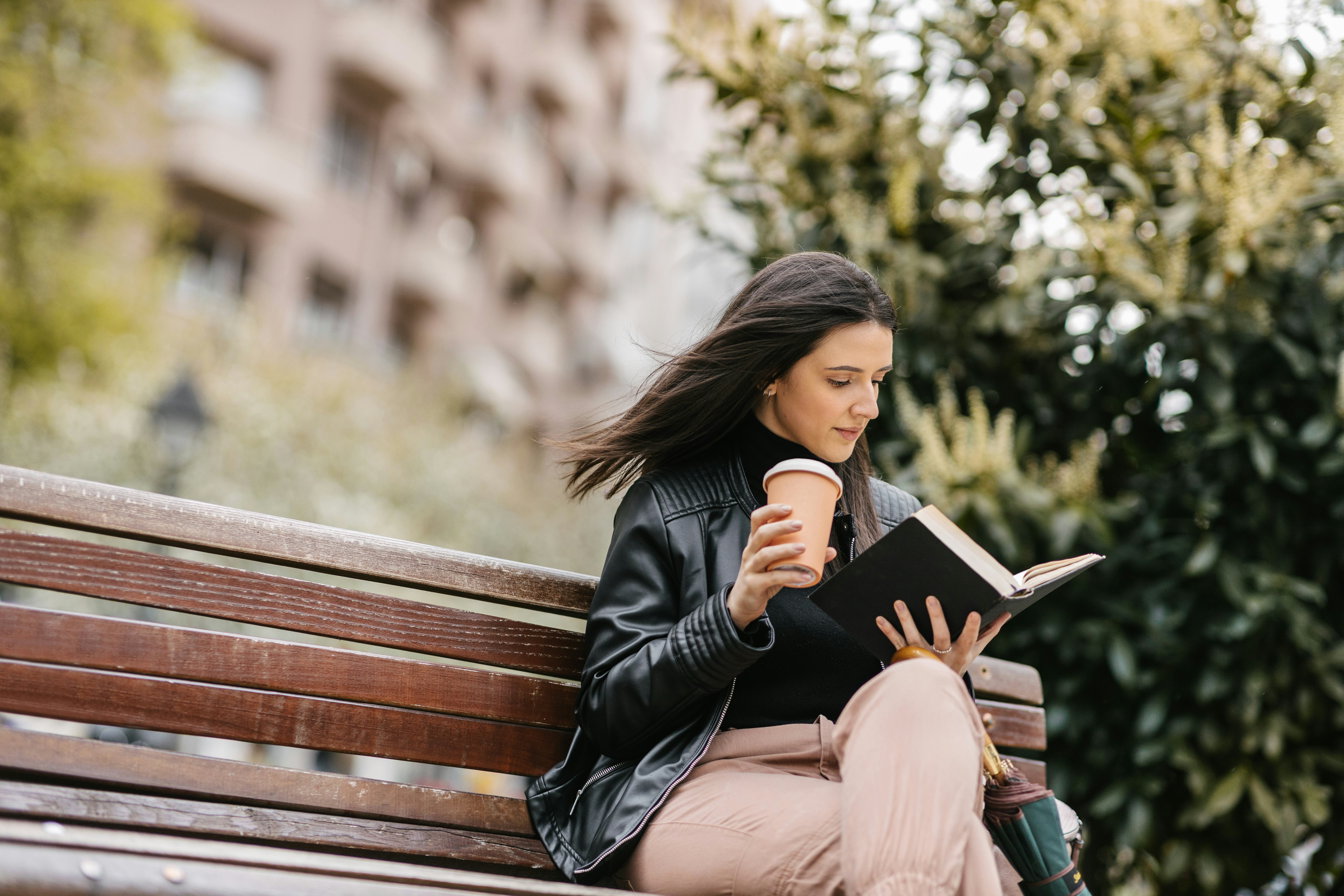 A young female student sitting in a park on a bench and reading a book. Coffee and umbrella