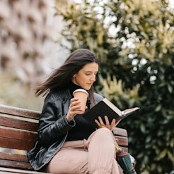 A young female student sitting in a park on a bench and reading a book. Coffee and umbrella