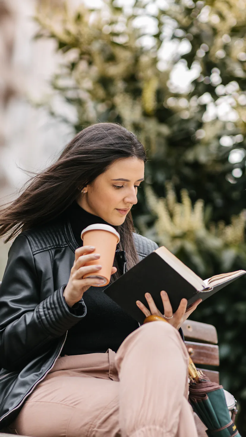 A young female student sitting in a park on a bench and reading a book. Coffee and umbrella