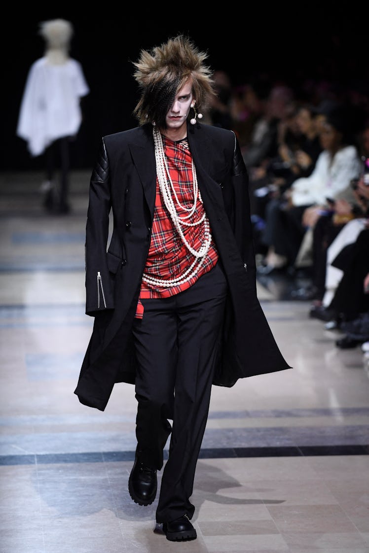 Male model in Junya Watanabe checkered red shirt with long pearl necklace, black coat & pants at Par...