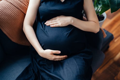 Cropped shot of Asian pregnant woman gently touching her baby bump, relaxing on the sofa at home. Mo...