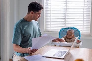 A man holding a paper and working on his laptop at a table while his baby sits beside him in a high ...