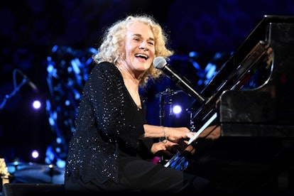 Carole King playing the piano and singing, wearing a black jacket.