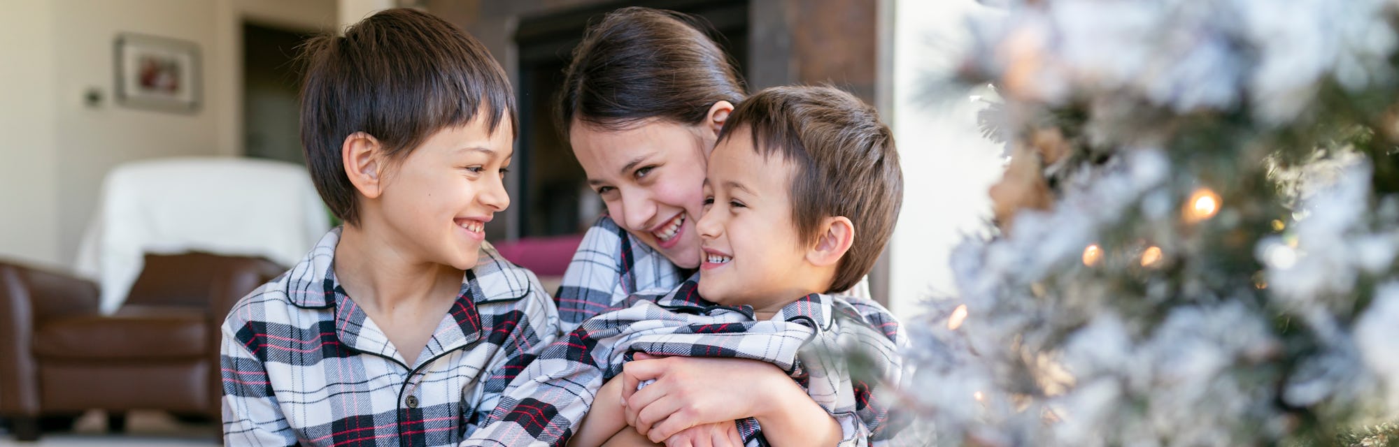 Three siblings wearing matching holiday pajamas sitting near a Christmas tree.