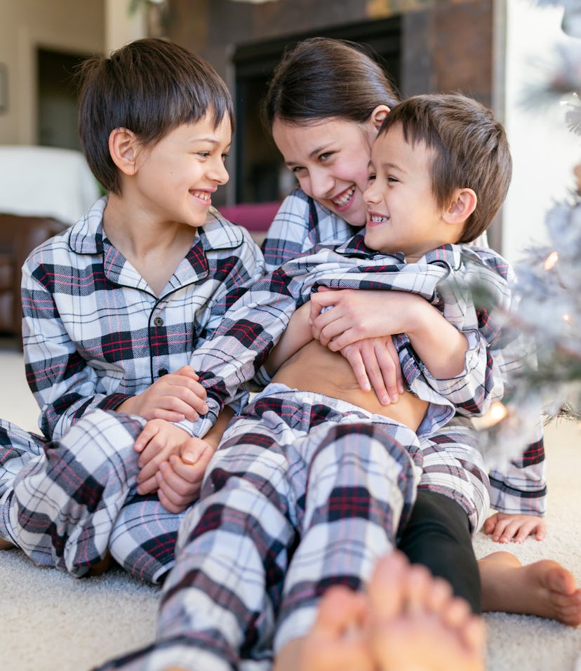 Three siblings wearing matching holiday pajamas sitting near a Christmas tree.
