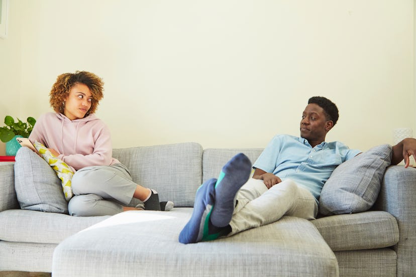 Young couple sitting apart on the sofa. They are looking at each other with irritated expressions. i...